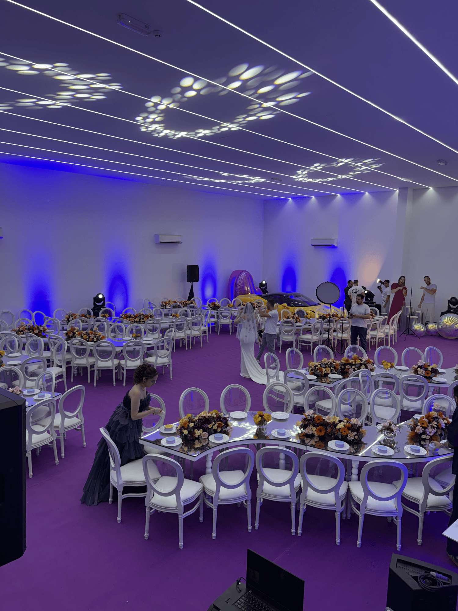 Wedding reception setup with floral table arrangements, white chairs and violet lighting